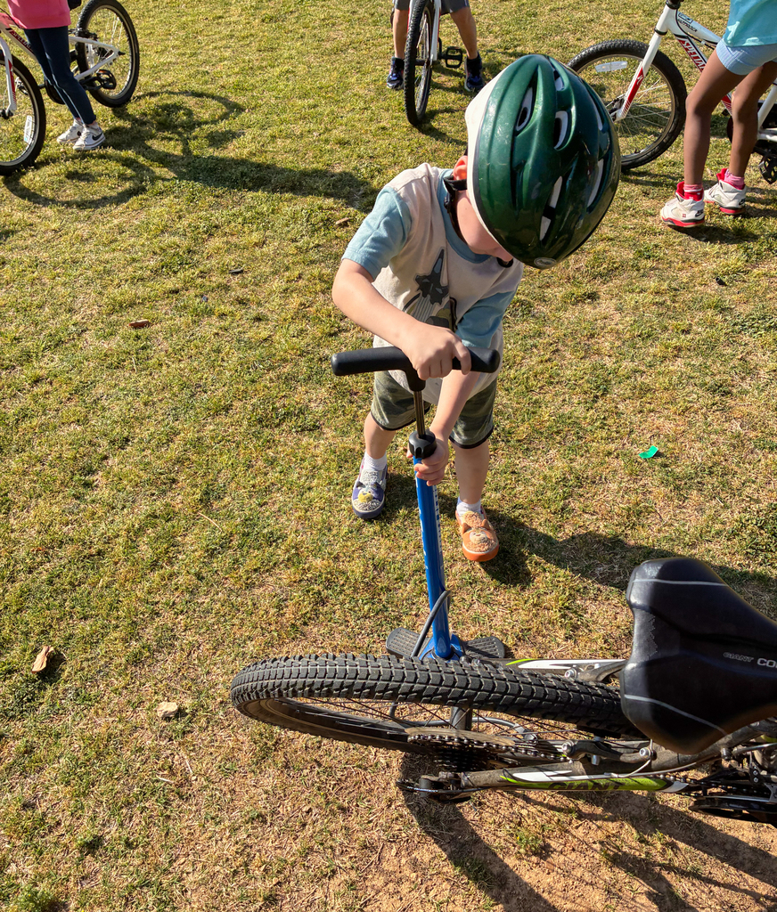 A child in a helmet is pushing a bike on grass. Other children ride bicycles in the background.