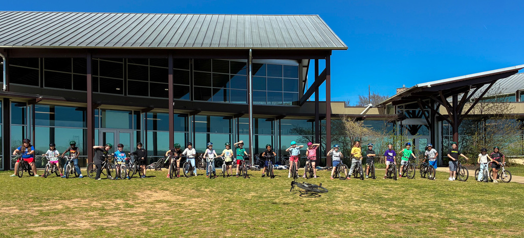 A group stands in a grassy area with bicycles in front of a modern building.