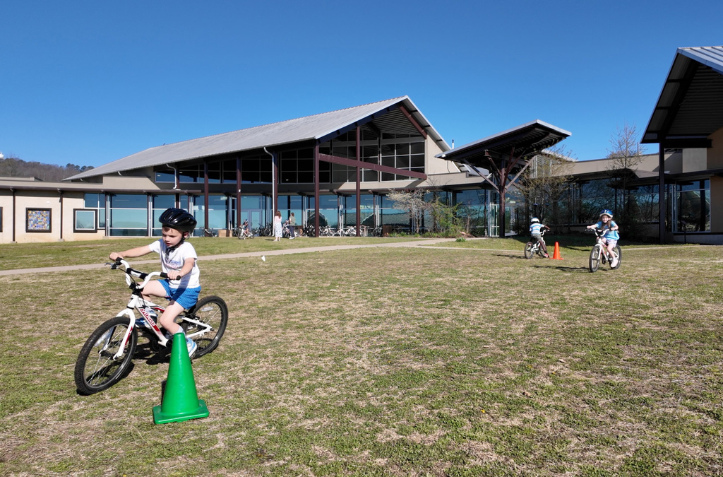 Children ride bicycles in an outdoor field with a building in the background. One child maneuvers through green cones.
