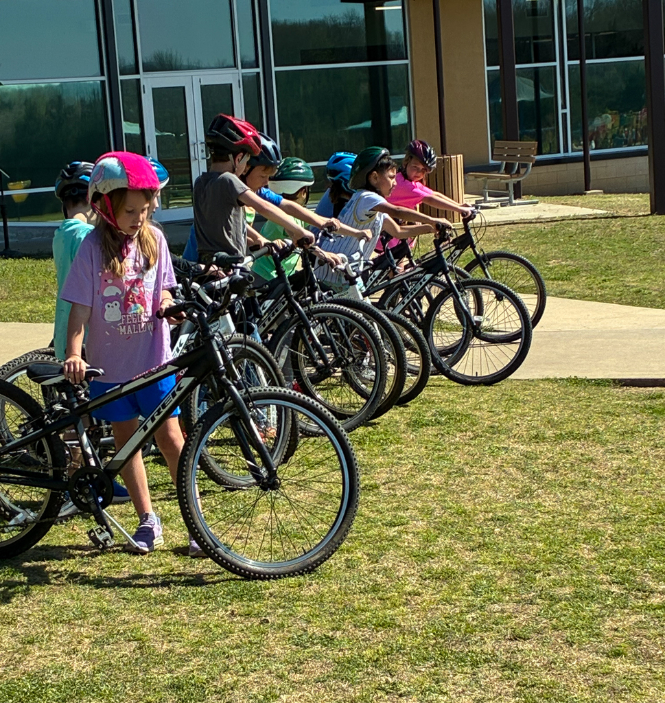 Children in helmets line up with bicycles on a grass field in front of a building.
