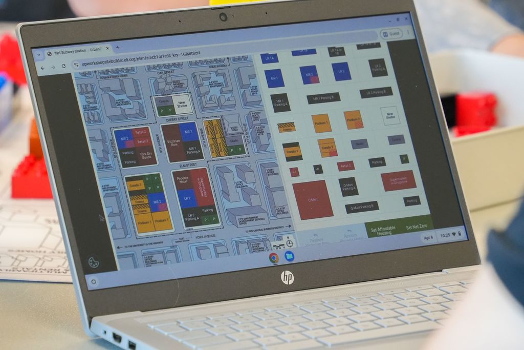 A laptop open on a desk, displaying a floor plan with various colored blocks. Behind, a person in a blue shirt.