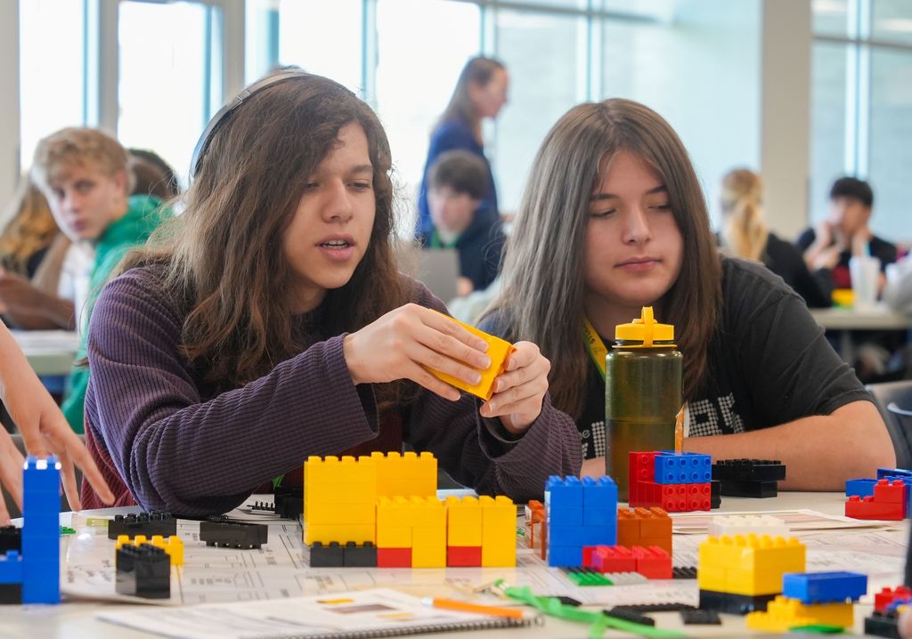 Two girls work on a colorful Lego project, surrounded by other students in a room with large windows.