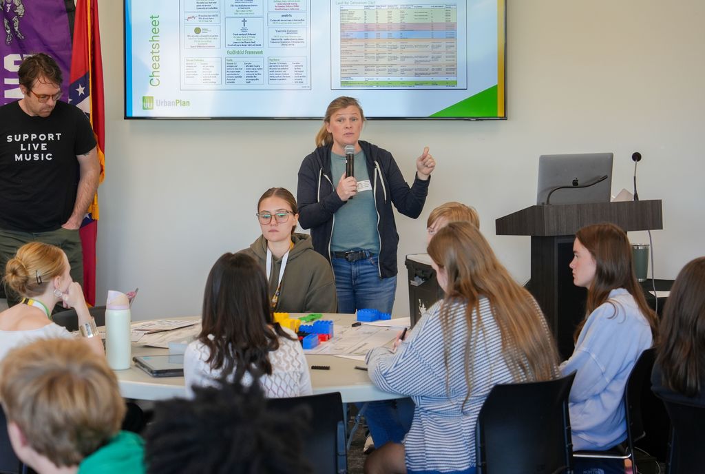 Woman stands at a table, speaking to a group of young adults. A projector screen shows a graph.