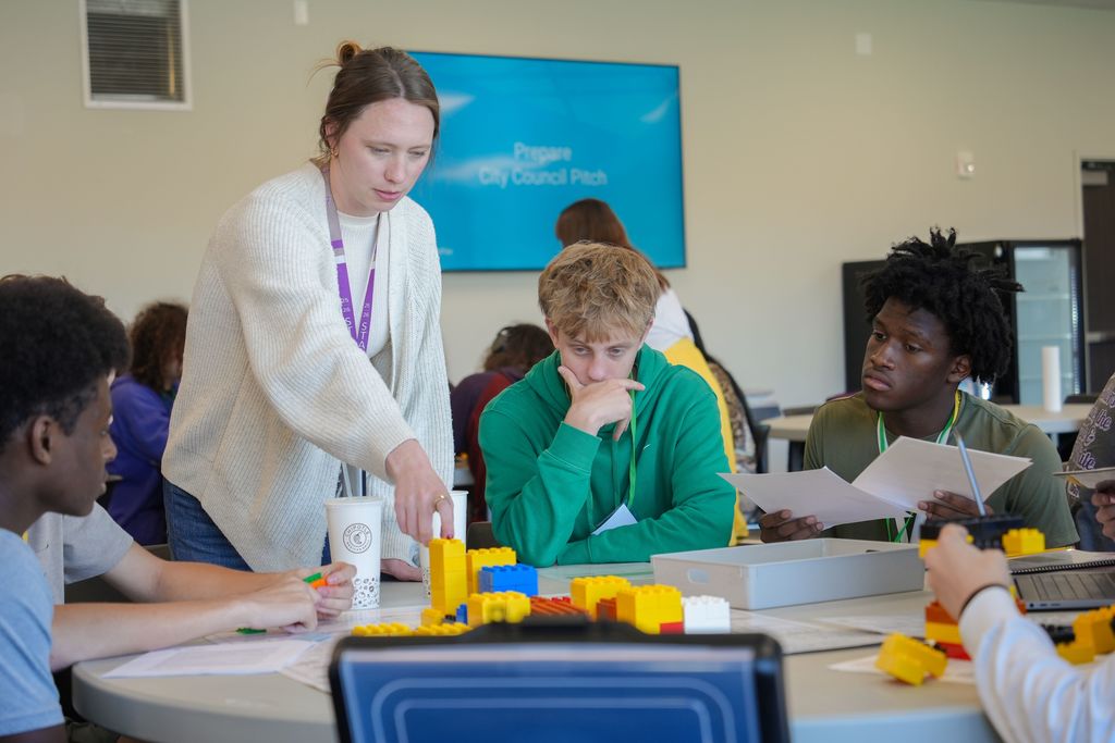 A woman stands over a table with blocks as several young people sit around it, looking at papers.
