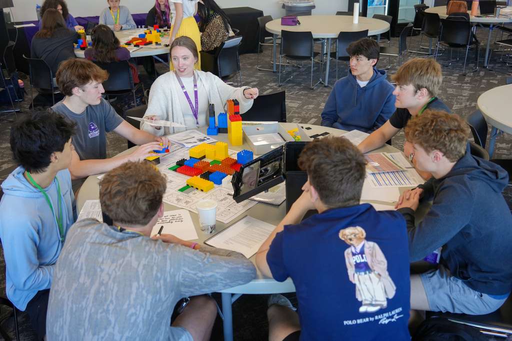 Students gather around a table in a classroom, constructing something with blocks. Some are focused on a computer screen.