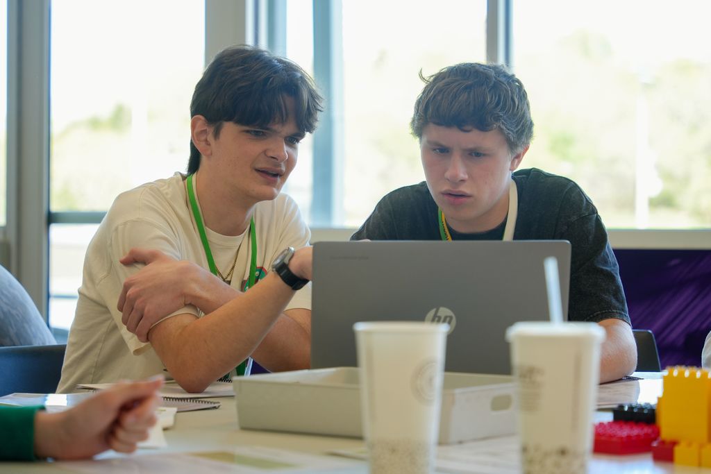 Two young men work together at a table, one using a laptop, the other gesturing. Cups and building blocks are on the table.