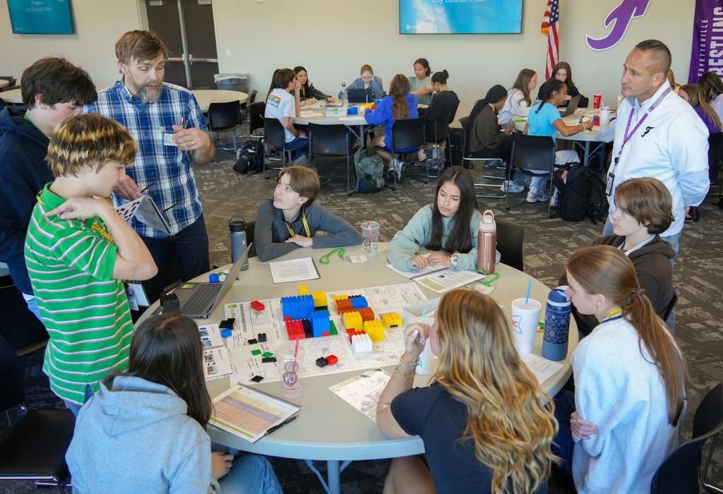 A group of students of various ages gathered around a table with building blocks, guided by a male instructor.
