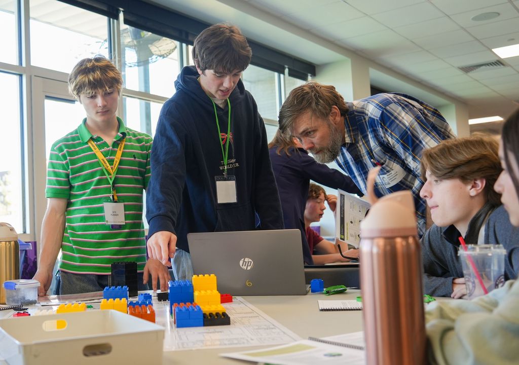 Five students work on a project using Legos and a laptop on a table. One student leans over another.