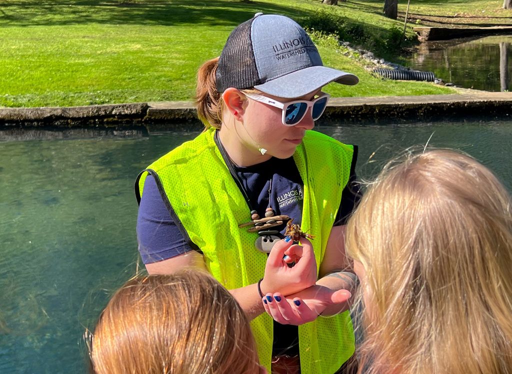 A person in a neon green vest holds a small creature while three children look on.