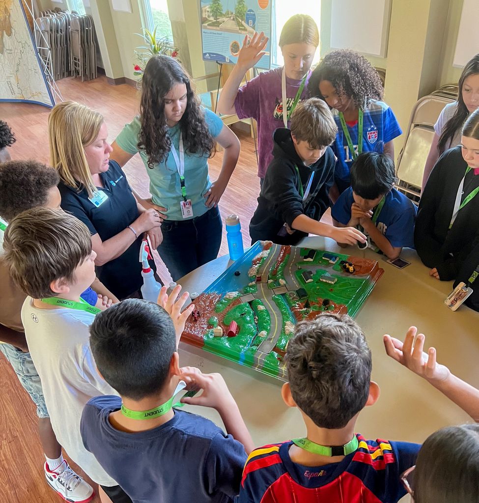 Several children and adults are gathered around a table with a model train set, likely in a classroom.