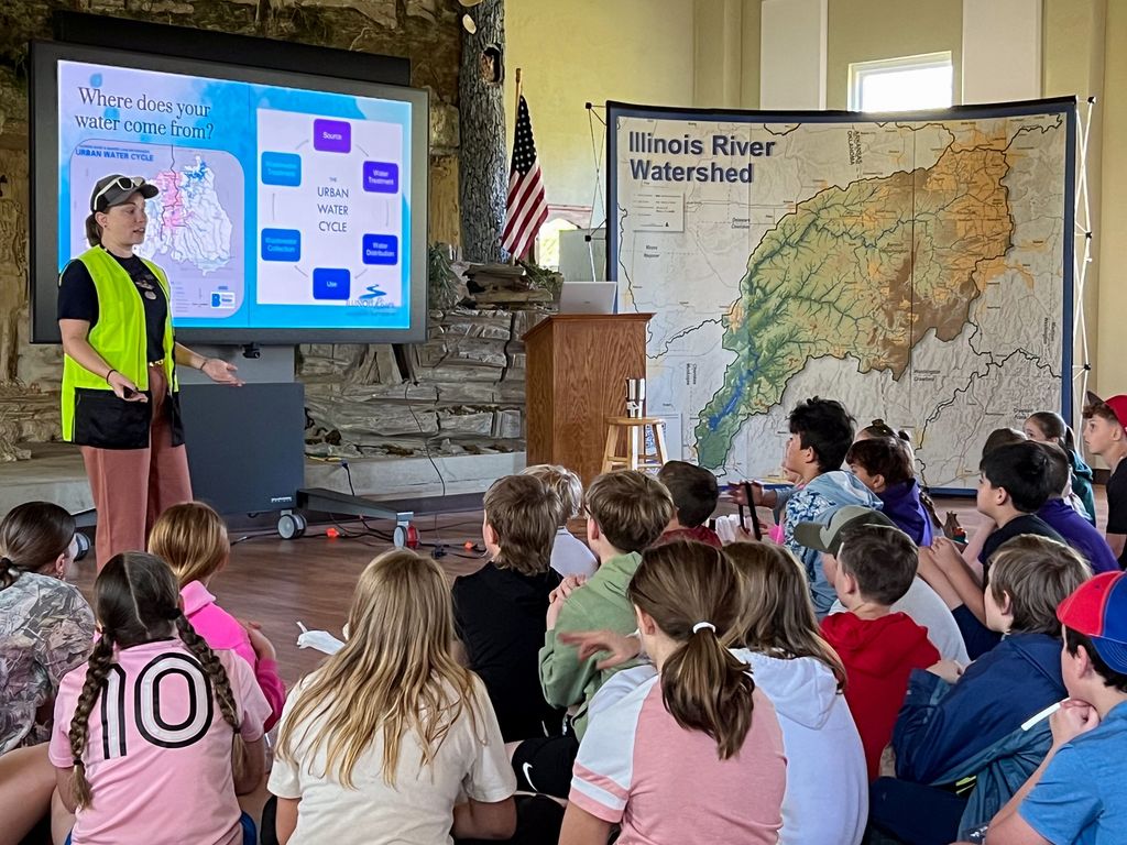Woman in safety vest speaks to students. Map of Illinois River Watershed displayed behind her.