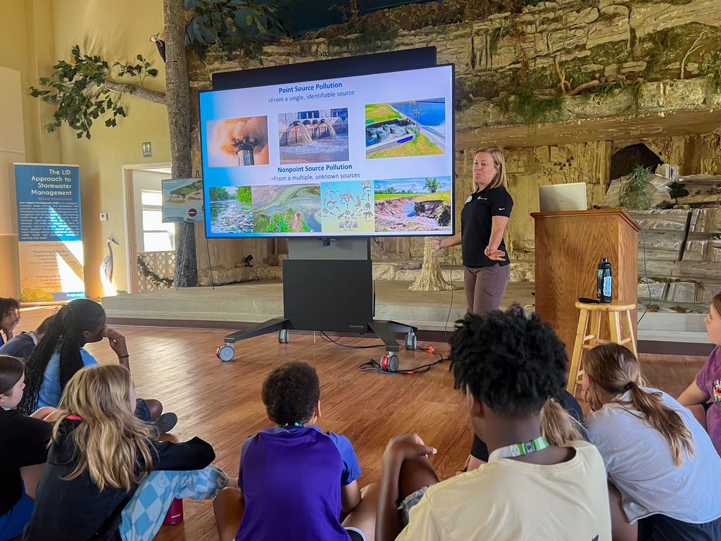 A woman speaks to a group of children around a circle in a classroom, using a projector screen.