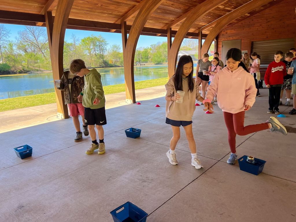 Children play a game with blue bins on a concrete floor. Behind them, a lake is visible.