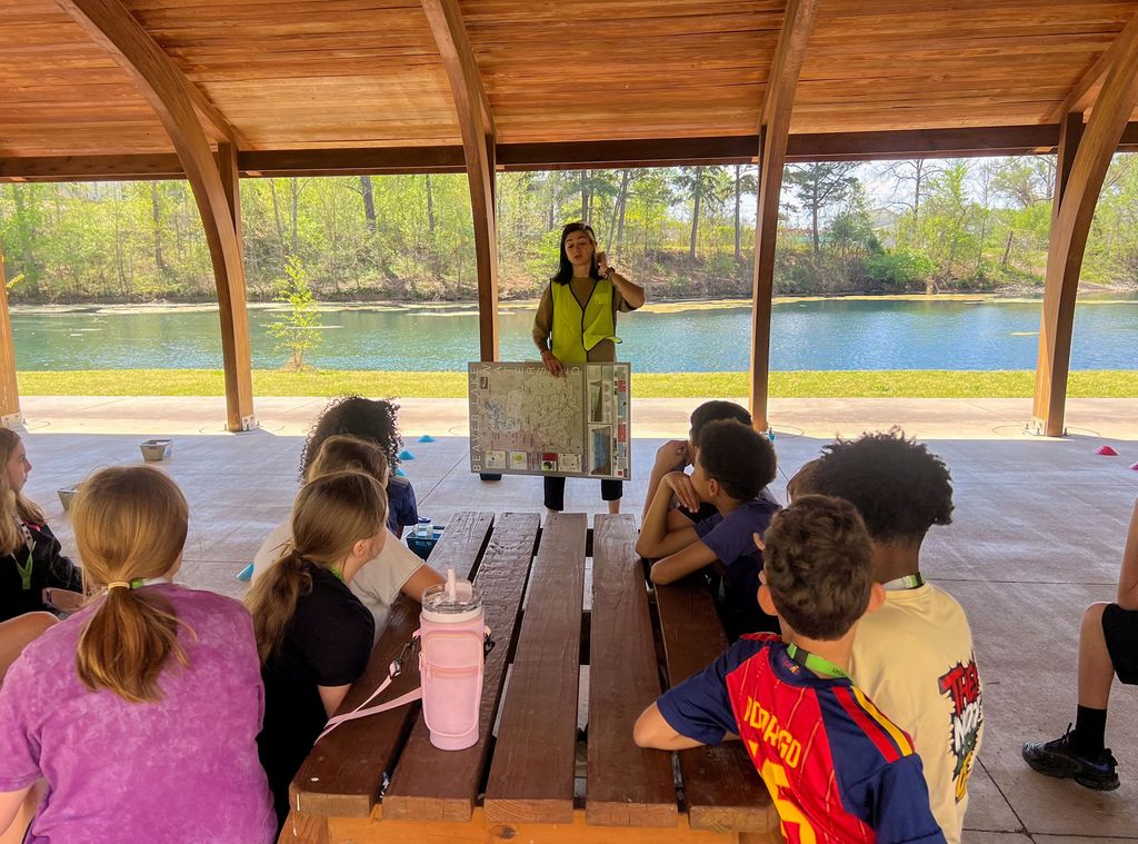 A woman stands in a gazebo, holding a board, with students seated around a picnic table, overlooking a lake.