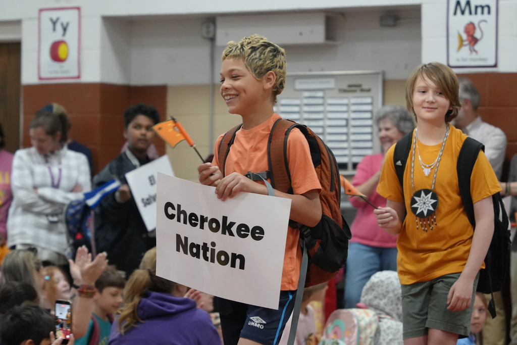 Two children hold a sign reading "Cherokee Nation." A crowd of people surrounds them in a hallway.