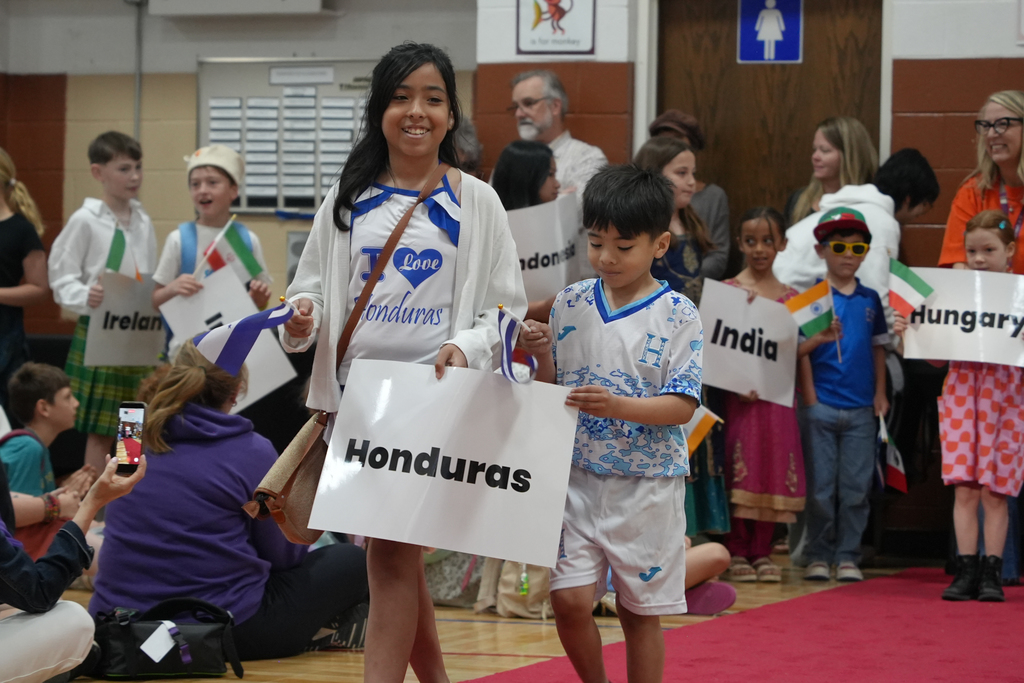 Kids in various national dress walk on a red carpet, one holding a sign that says "Honduras".