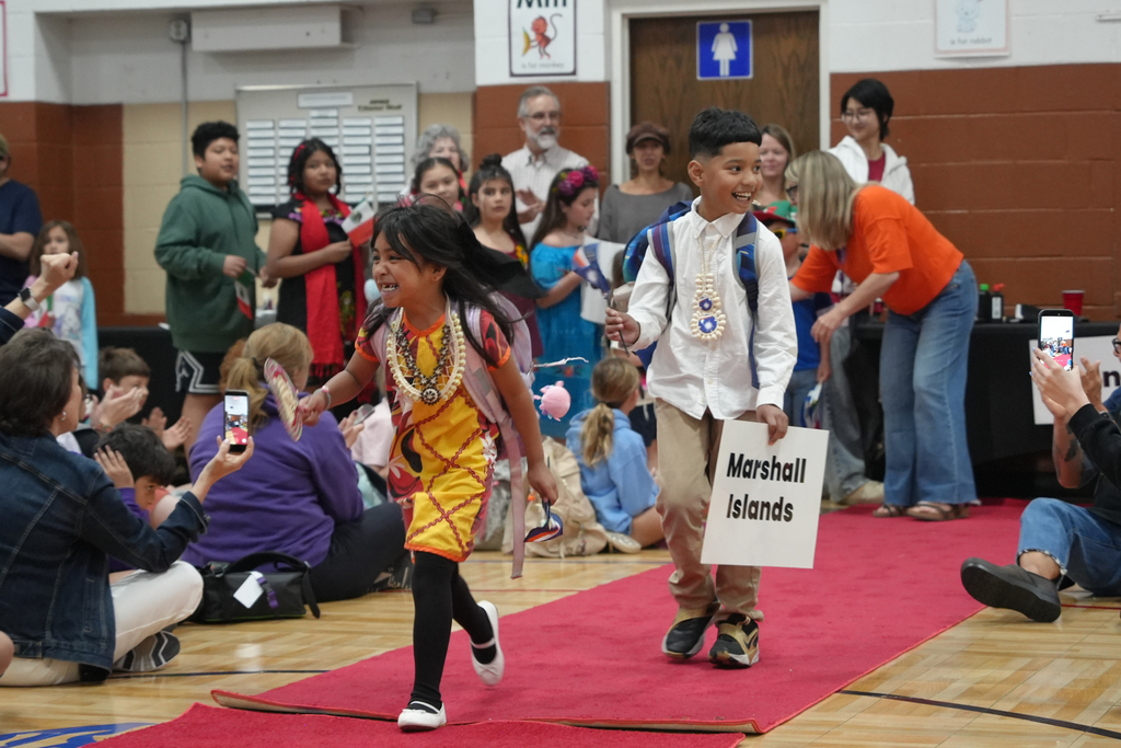 Two children walking on a red carpet, holding signs. Behind them, a crowd of people watches, some standing and some sitting.