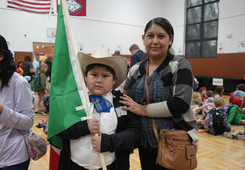 A woman stands next to a child holding a flag. They are in a room with other people.