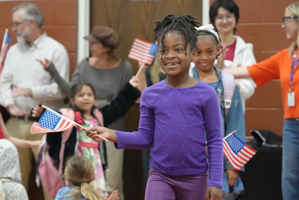 A young girl in purple waves two flags while other children and adults stand behind her.