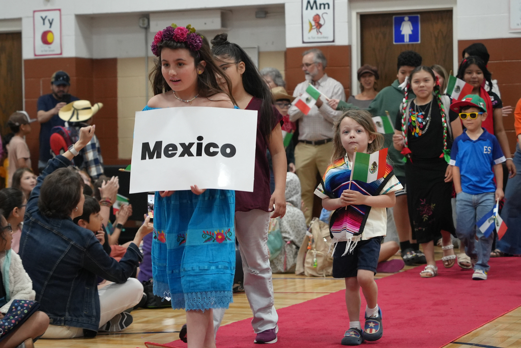 A girl holds a sign saying Mexico while walking on a red carpet. Behind her, a young child carries a flag.