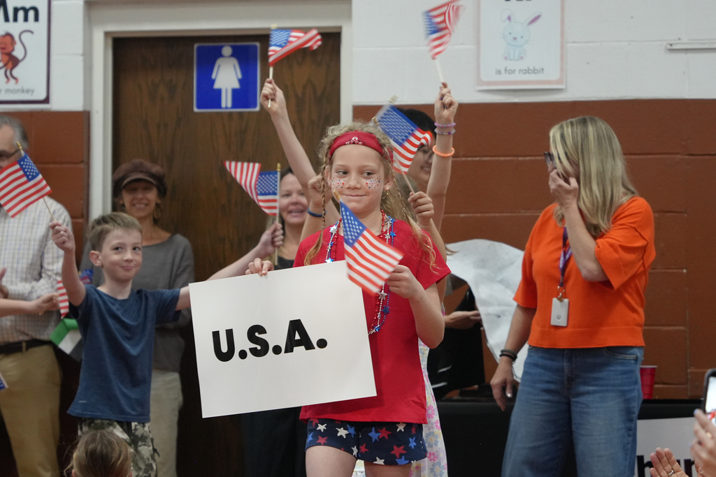 Children and adults hold small flags and a sign that says U.S.A. They stand in a room with a door and brown walls.