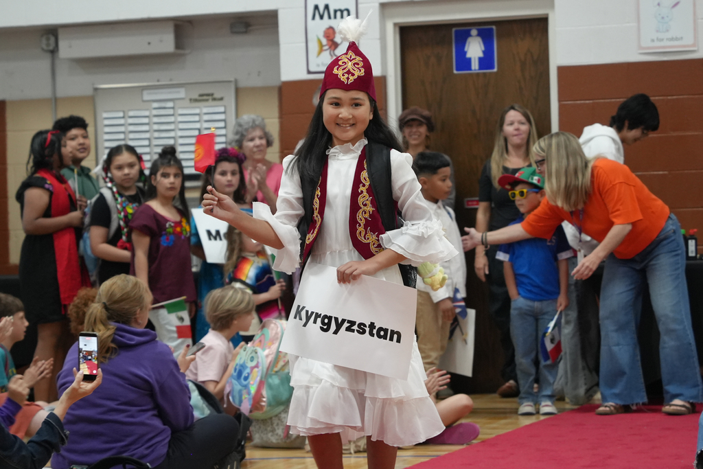 Person in a white dress with a red hat walks on a red carpet, displaying a white sign with the text "Kyrgyzstan".