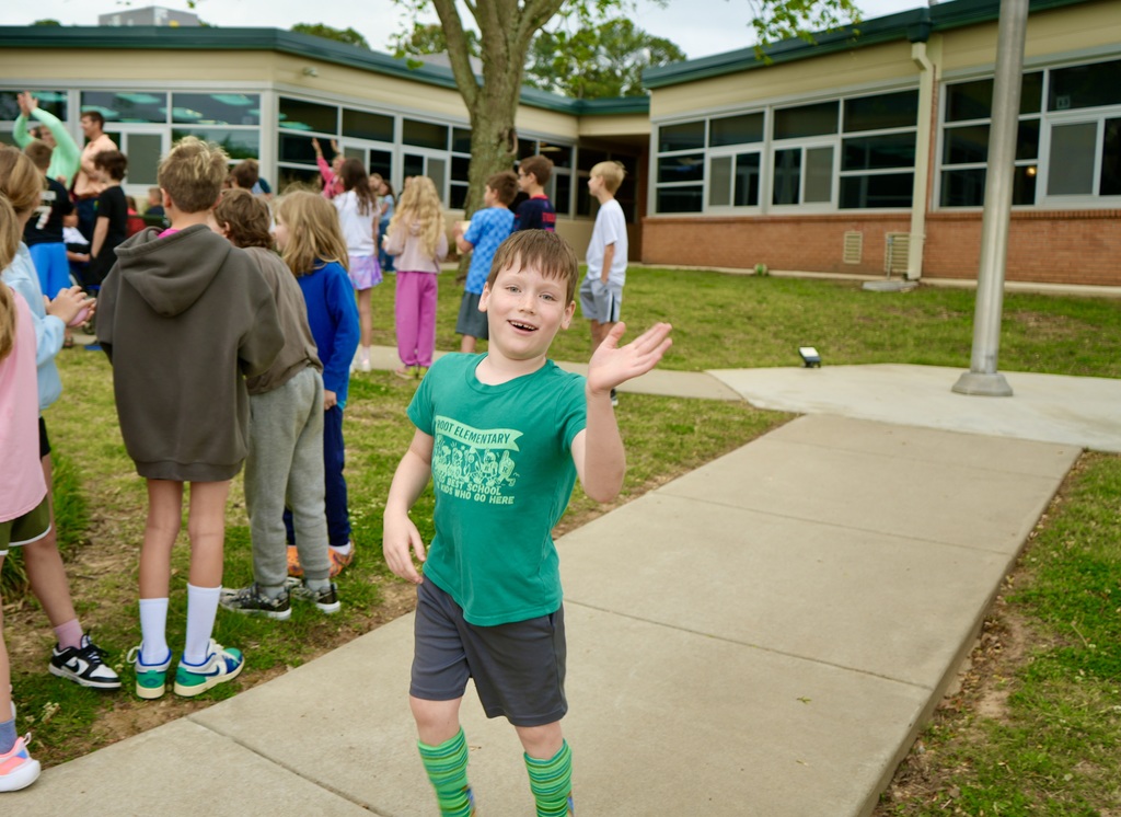 A young boy wearing a green t-shirt stands on a sidewalk in front of a building, waving.