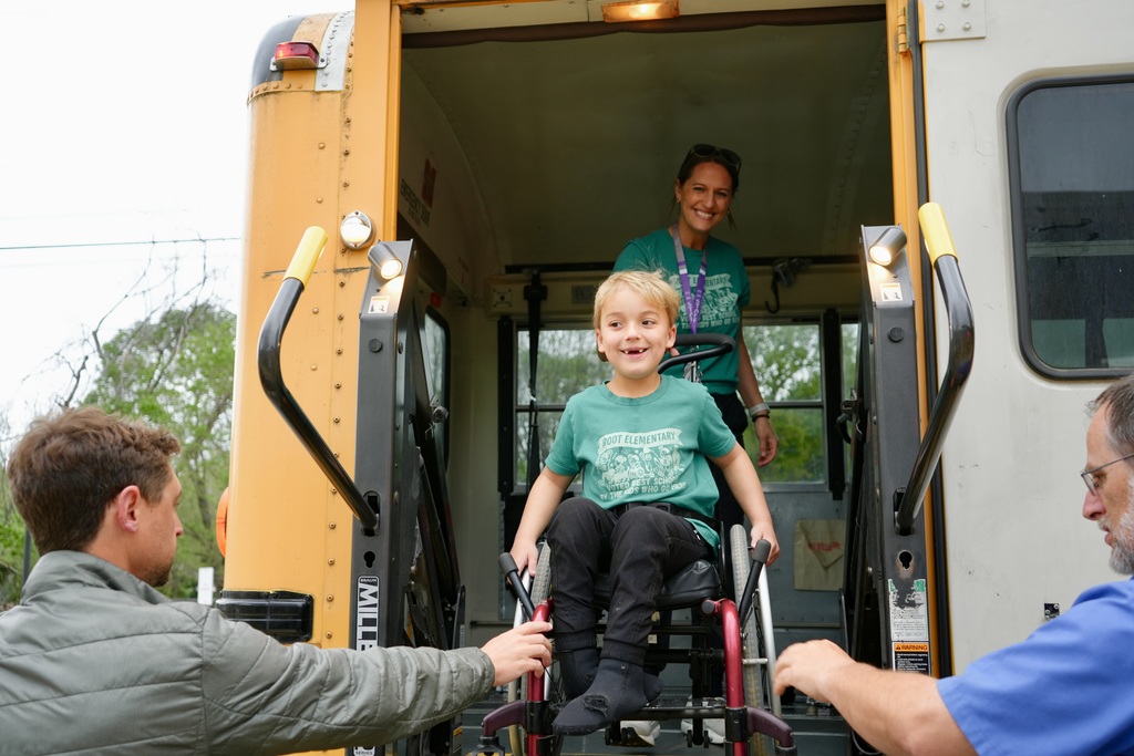 A boy in a wheelchair is boarding a bus with assistance from two men. The woman is standing near the open door.
