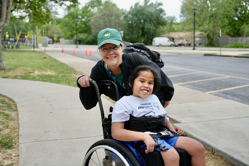 A woman wearing a green cap and glasses pushes a young girl in a wheelchair on a sidewalk.