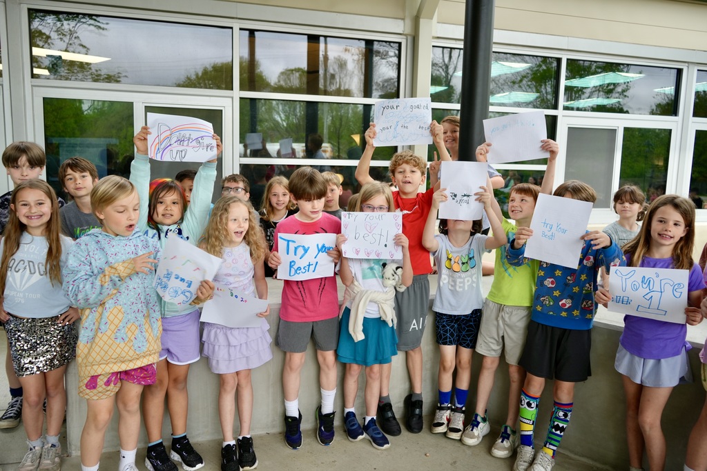 A group of children in various clothing holding up signs in front of a building with glass walls.