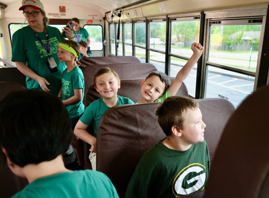 Kids wearing green shirts sit on a bus with two adults; one child has a yellow headband.