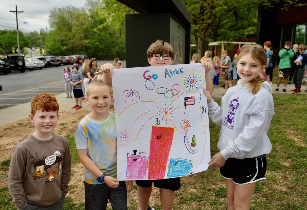 Four kids hold a drawing of a firework and an Olympic flame. They stand in front of a building.