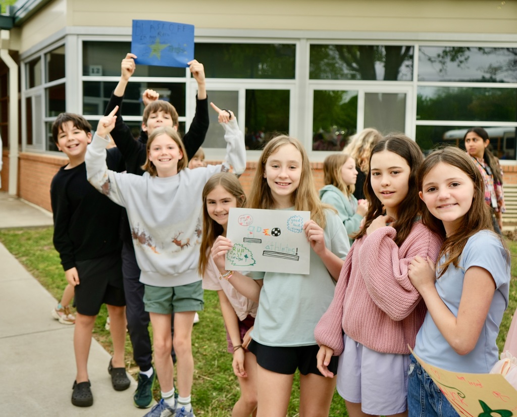 Kids stand on a sidewalk holding signs, one child holds a blue paper. They smile, and some wear sneakers. Behind them is a building with glass windows.
