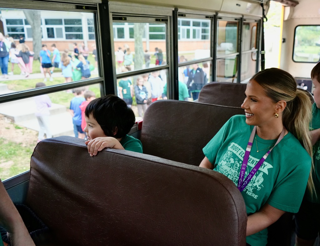 People sit inside a bus, one woman smiles while looking forward, a child rests their hand on the seat, and a building is visible outside.