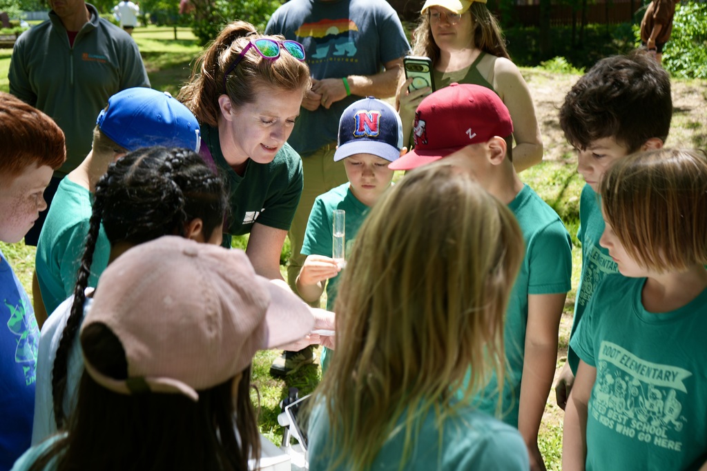 A group of people, likely students, in green shirts, standing in a park. A woman is teaching the students.