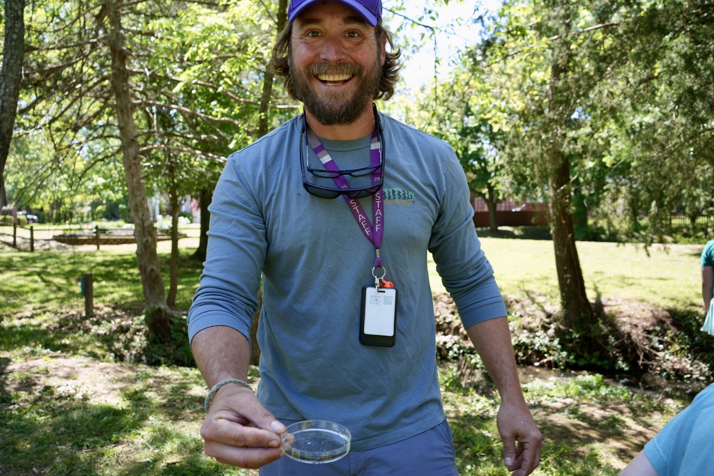 A man wearing a blue shirt and purple lanyard smiles and holds a clear petri dish outdoors.