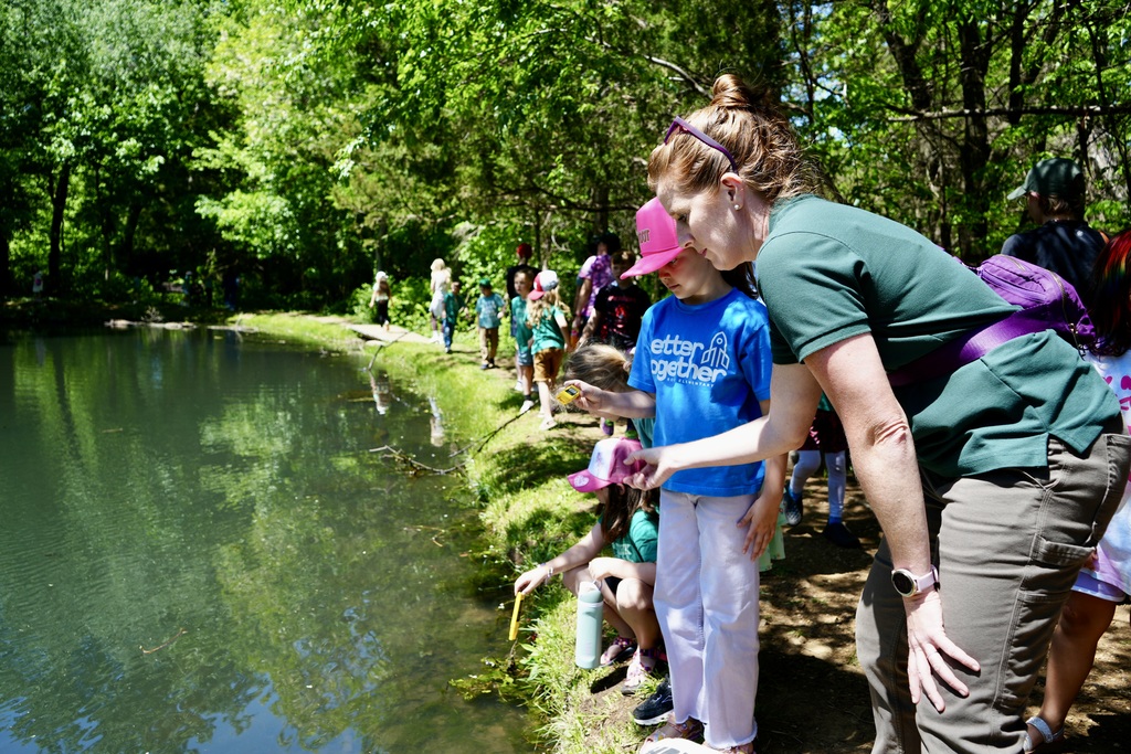 A woman teaching a young girl and others how to catch fish in a pond, surrounded by trees.