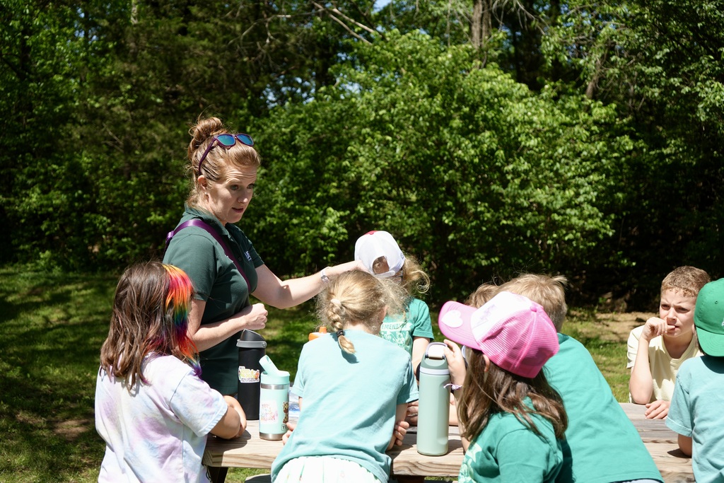 A woman in green assists a group of children wearing green shirts and hats, seated around a table in a wooded area.