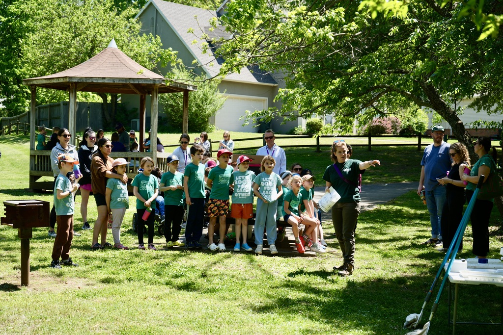A group of children and adults in green shirts stands in a park under a gazebo. Some are wearing hats.