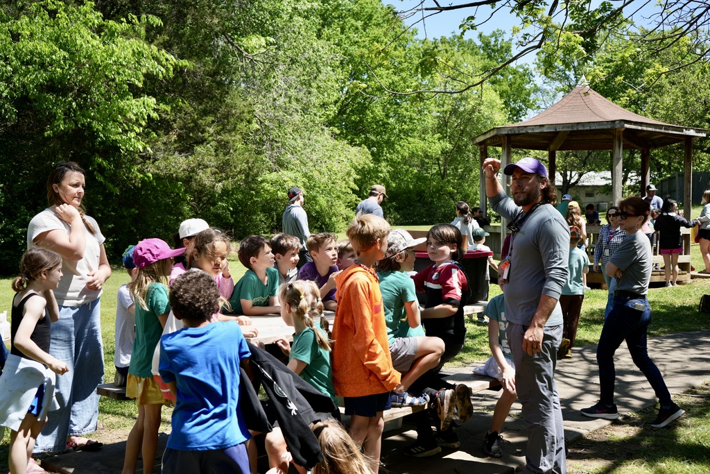 People gather around a picnic table in a park, with a gazebo in the background. Some wear hats.