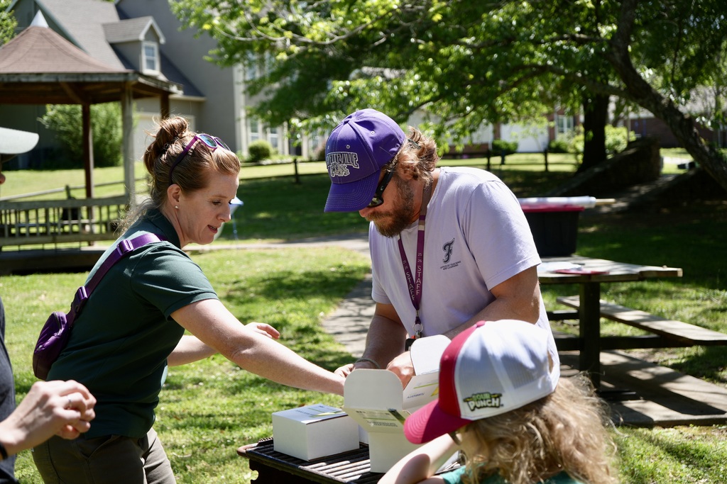 Woman in green shirt holds box as man in white shirt and purple cap looks inside. Child in pink cap watches. Picnic tables and gazebo in background.