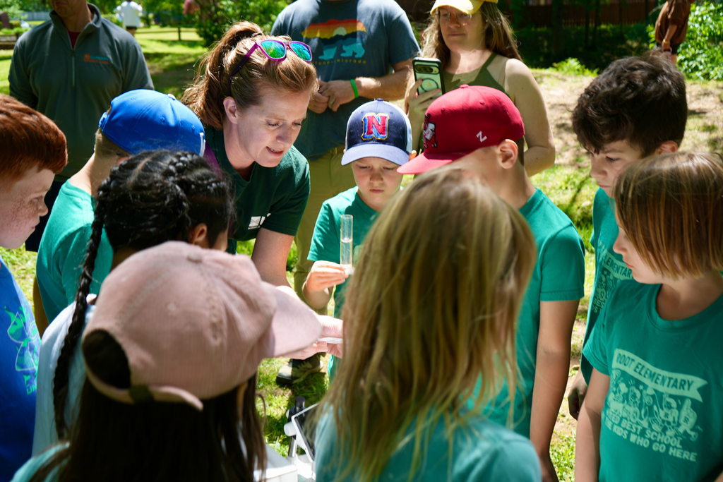 People outdoors. A woman shows a group of children a sample in a tube.