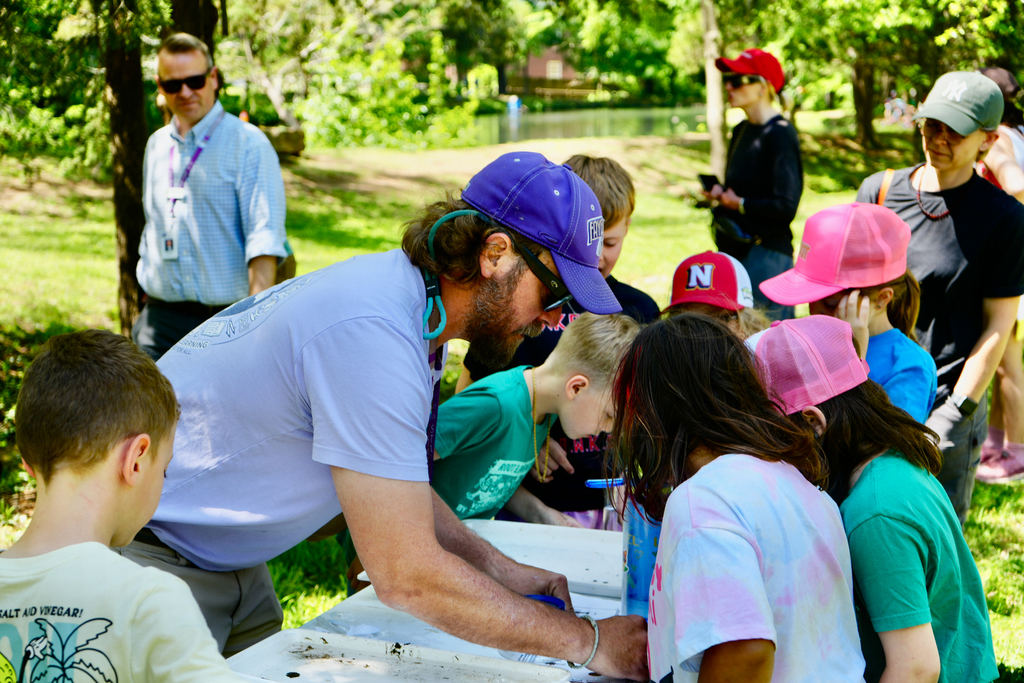 A group of children and adults gather around a table outdoors, with a person in a blue hat showing something to the children.