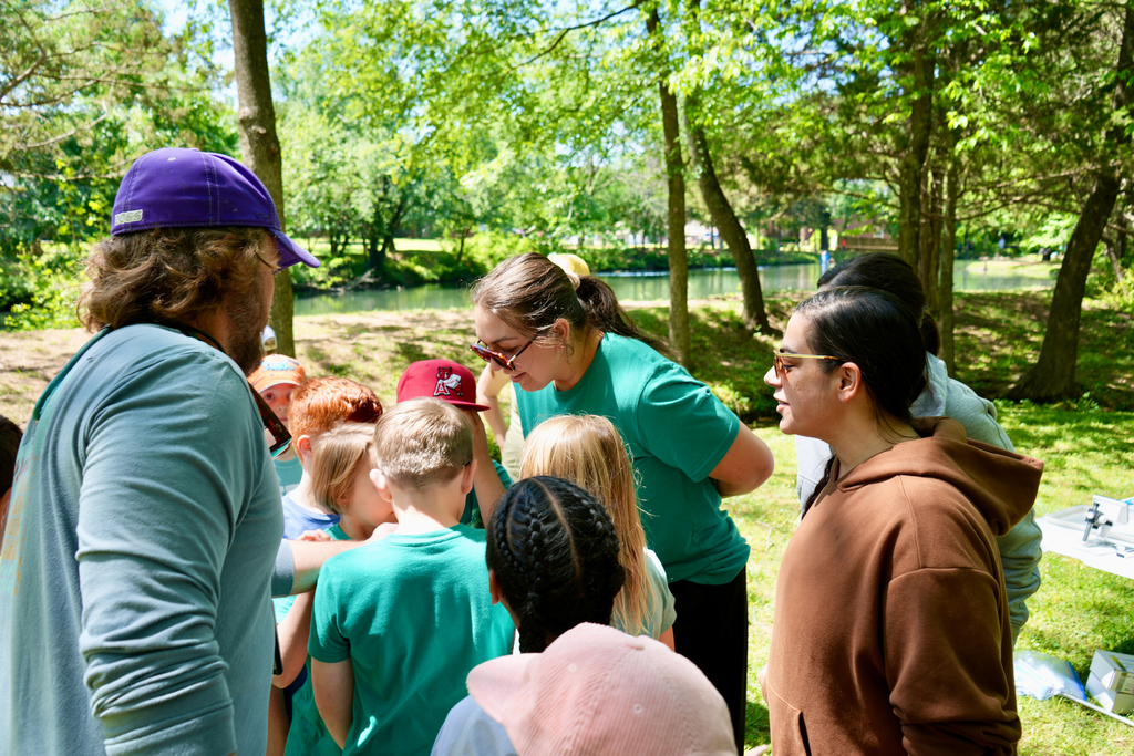 A group of children and adults wearing green shirts gather in a park with a river in the background.