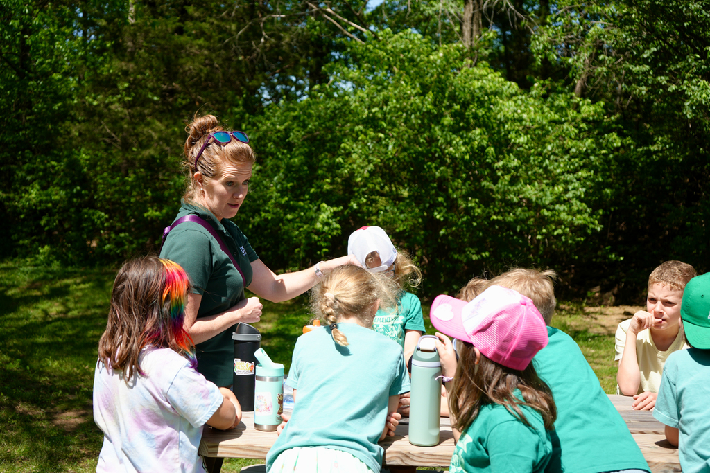 A woman helps children at a picnic table. They sit in a circle, some wear hats. Trees are in the background.