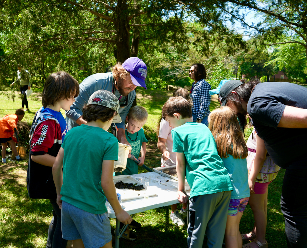 Group of children and adults gathered around a table in a park, examining items on the table.