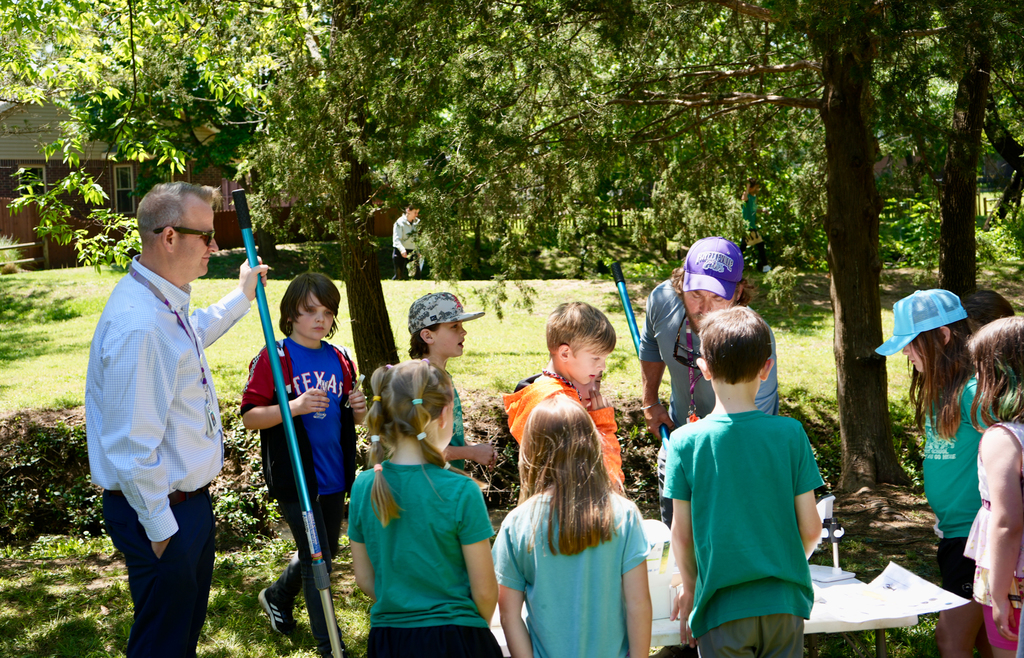 A man in a white shirt instructs a group of children in green shirts under a canopy of trees.
