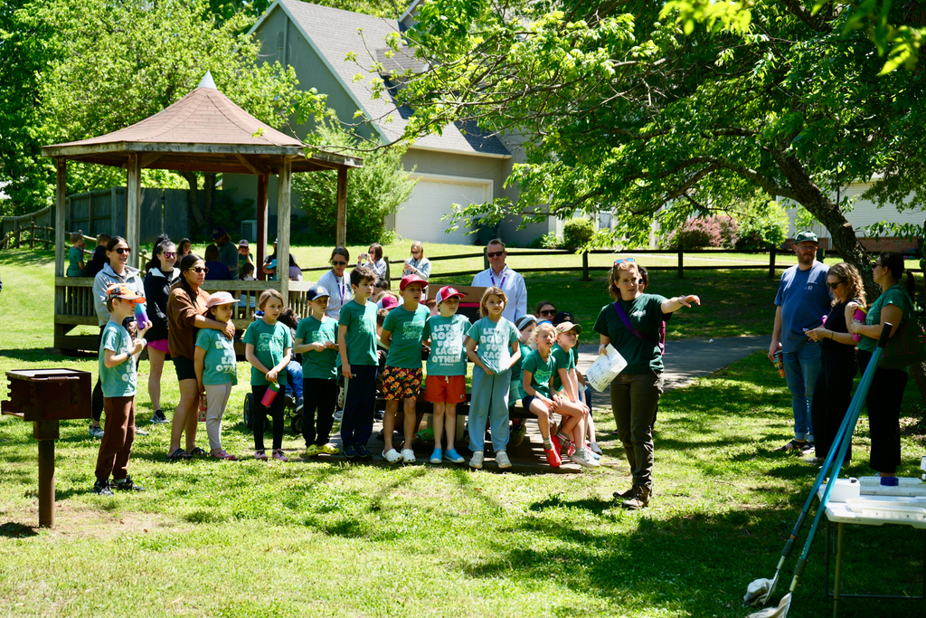 A group of people in green shirts, some wearing hats, are standing on a grassy area. A person stands in front, speaking. Behind them, a gazebo, a house, and trees.