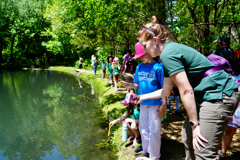 A woman and children are looking into a pond, observing fish. The woman wears a green shirt and sunglasses.