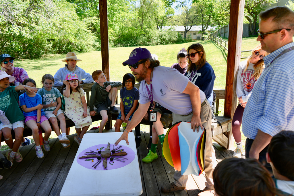 A man demonstrates a craft to a group of children seated on benches under a gazebo.